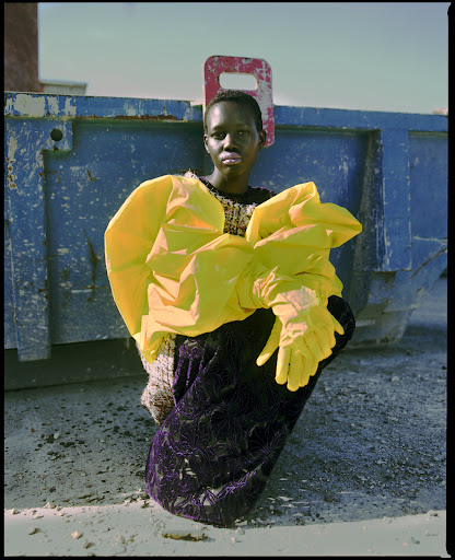 Fashion photo featuring a Black woman in an avant-garde yellow ensemble, posed against a blue dumpster under the open sky.