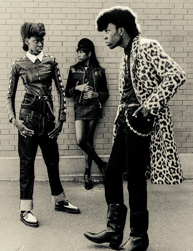 Black and white photo of three adult women in stylish 1980s-inspired clothing posing against a brick wall outdoors on a city street.