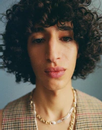Close-up portrait of a man with curly dark hair wearing a plaid vest and layered chain necklaces against a solid background