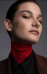 Close-up portrait of a woman wearing a high red turtleneck under a black collar and brown blazer, against a grey background.