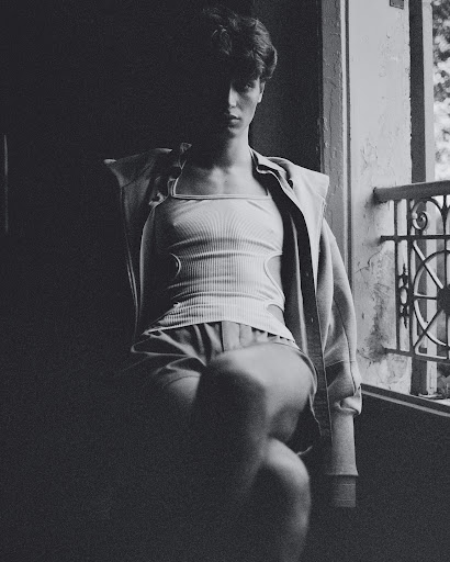 Monochrome portrait of a young man seated near a balcony window in Paris, featuring makeup by Camille Lutz.