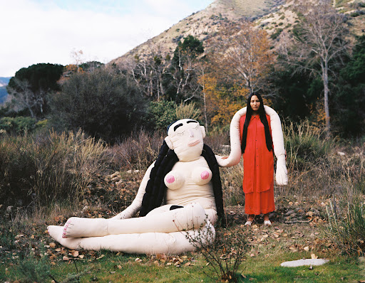 Artistic outdoor portrait of a woman in a red dress with a large stuffed figure on a grassy field with a mountain background.