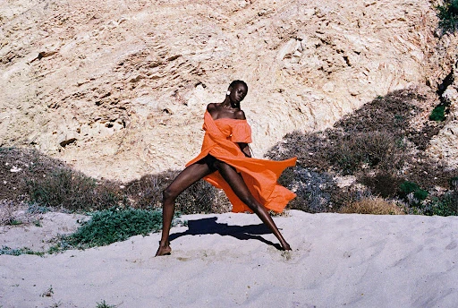 Woman in orange dress poses on sand in front of rock formation; fashion, outdoor setting, sandy ground, flowing fabric, off-shoulder style.