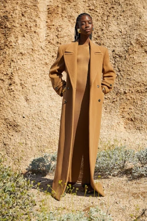 Black model with braided hair wearing a long brown wool coat and matching dress standing against a sandy rocky hillside.