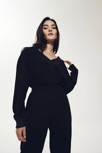 Portrait of a woman in a black jumpsuit posing against a neutral background, looking at the camera with a neutral expression.