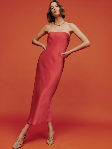Model in a strapless pink midi dress posing against a warm orange studio backdrop with makeup by Hadia Kabir
