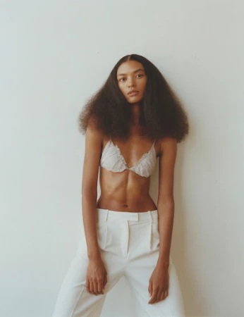 Model with voluminous dark hair wearing a light bralette and white trousers against a neutral wall in Los Angeles.