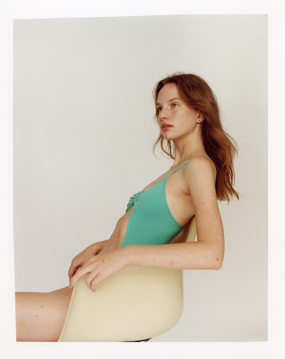 Profile portrait of a woman with red hair wearing a teal swimsuit seated on a light yellow chair against a plain white background