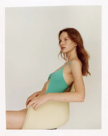 Profile portrait of a woman with red hair wearing a teal swimsuit seated on a light yellow chair against a plain white background