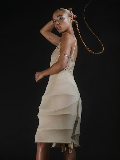 A fair-skinned woman wearing a beige dress and a blonde braid poses in a photo studio with a dark background.