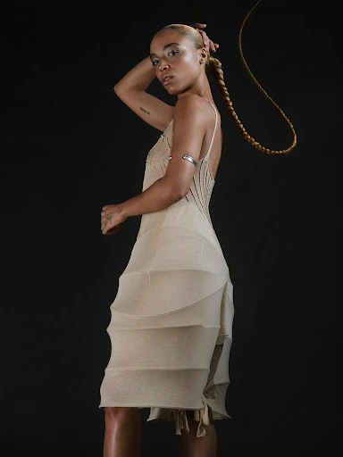 A fair-skinned woman wearing a beige dress and a blonde braid poses in a photo studio with a dark background.