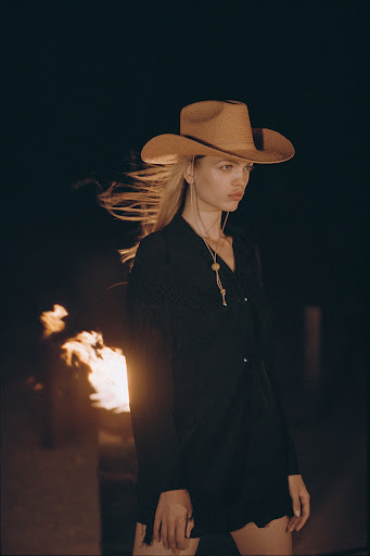 Young woman with blonde hair and cowboy hat poses in black outfit outdoors by campfire at night, creating a fashion editorial image.