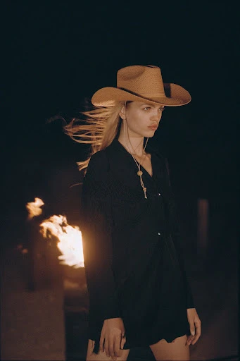 Young woman with blonde hair and cowboy hat poses in black outfit outdoors by campfire at night, creating a fashion editorial image.