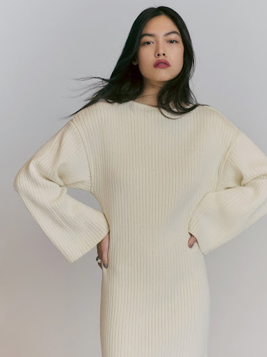Adult female wearing cream-colored knit dress stands in a photo studio with hands on hips; low-key fashion shot with soft lighting.