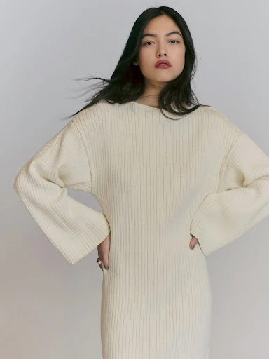 Adult female wearing cream-colored knit dress stands in a photo studio with hands on hips; low-key fashion shot with soft lighting.