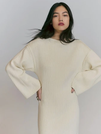 Adult female wearing cream-colored knit dress stands in a photo studio with hands on hips; low-key fashion shot with soft lighting.