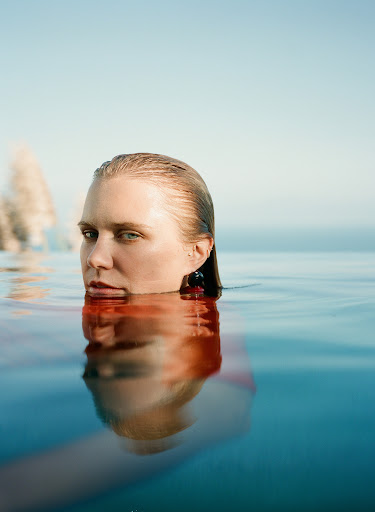 Woman in water up to her neck, with blue sky in the background. Her hair is wet and slicked back, and she is wearing earrings.