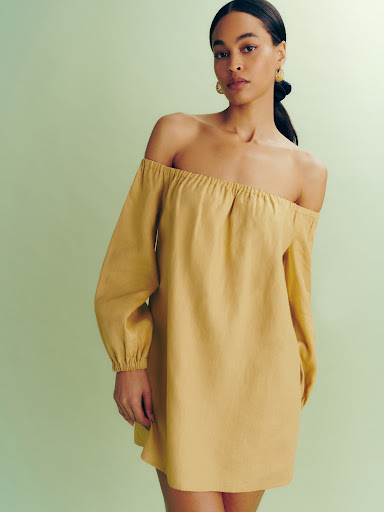 Brunette woman posing in a studio wearing a yellow off-the-shoulder dress in a studio setting with a light green background. Fashion editorial.