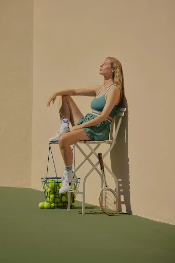 Female athlete sitting on chair with tennis balls and racquet in outdoor tennis setting, wearing a teal workout outfit, enjoying sun.