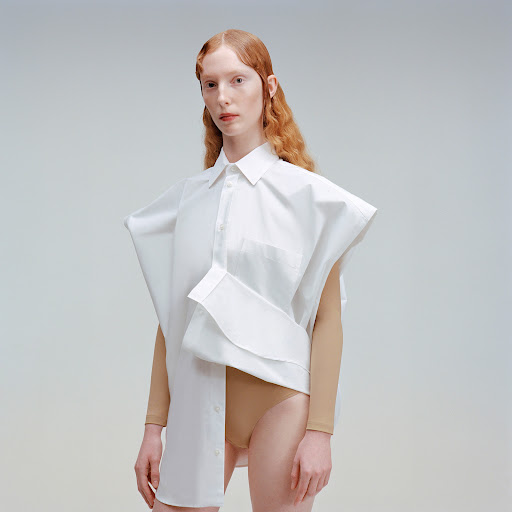 Fashion portrait of a red-haired woman wearing an oversized white shirt, posing in a photo studio with soft, neutral lighting.