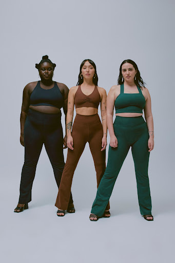 Three women pose in matching athletic wear against a neutral backdrop, showcasing different body types and styles in a studio setting.