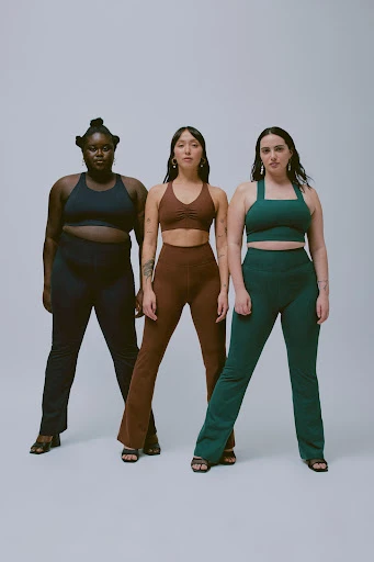 Three women pose in matching athletic wear against a neutral backdrop, showcasing different body types and styles in a studio setting.