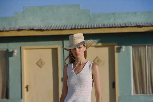Woman in a white dress and cowboy hat stands in front of a vintage-style building with light blue trim and a yellow door.