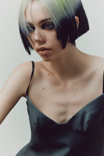 A portrait shot of a woman with long brown hair wearing a black top in front of a neutral background with soft lighting.