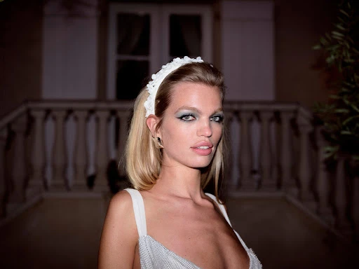 Portrait of a blonde woman wearing a white headband and winged eyeliner standing on a balcony at night.