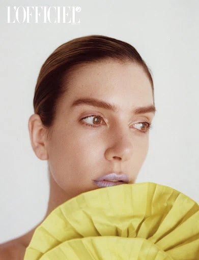 Close-up fashion portrait of an adult female model with slicked-back hair, lilac lipstick, and a yellow ruffled top against a white background.