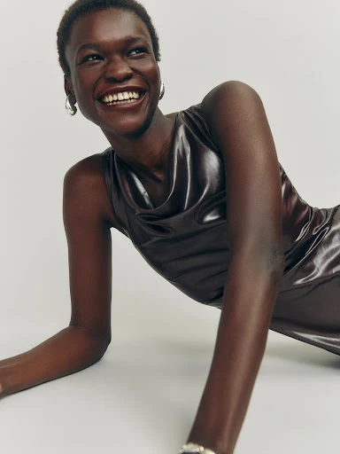 A smiling dark-skinned woman poses in a shiny brown dress against a neutral backdrop, showcasing fashion and beauty in a studio setting.