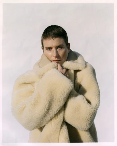Portrait of a woman with short hair wearing a fuzzy tan coat against a white background in a studio setting, fashion forward style.