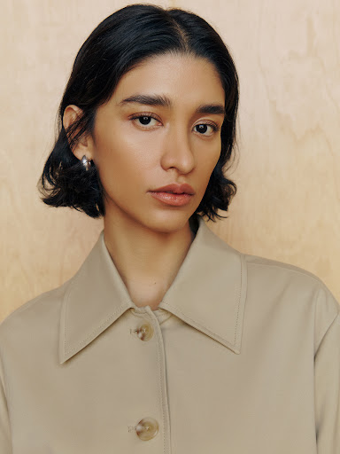 Portrait of an adult female with short black hair wearing a tan button-down shirt against a light brown, vertically-grained background.