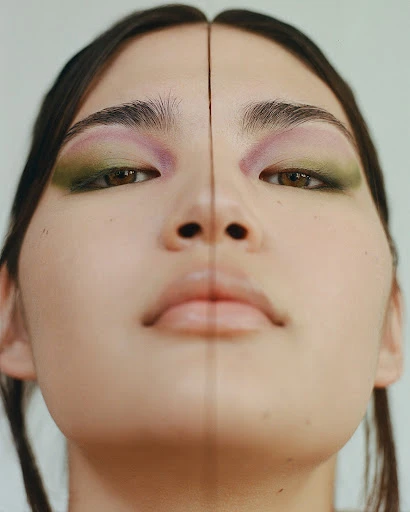 Close-up portrait of a young woman's face with colorful eyeshadow and makeup, split vertically with a slight mirrored effect.