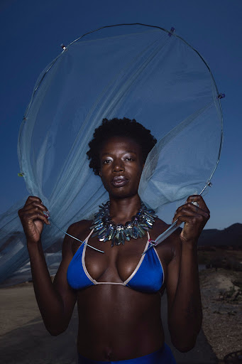 A woman in a blue bikini and jeweled necklace stands on a beach holding a transparent mesh material against a twilight sky.