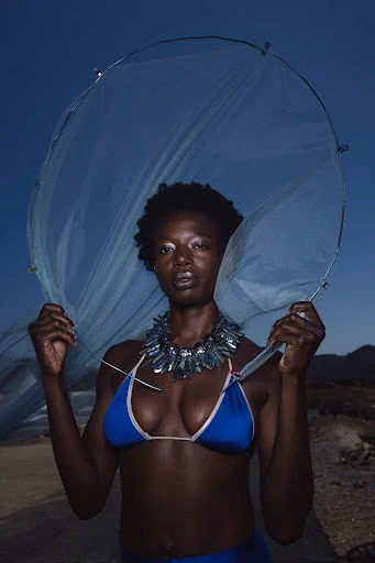 A woman in a blue bikini and jeweled necklace stands on a beach holding a transparent mesh material against a twilight sky.
