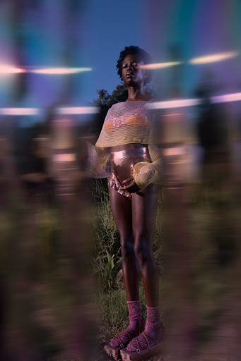 A lone Black woman stands in a field at dusk wearing avant-garde clothing, including a yellow netted top and gold ruffled shorts.