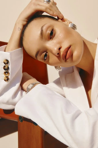 Close-up shot of a woman with silver hoop earrings wearing a white blazer leaning on a brown chair, bathed in soft, neutral light.