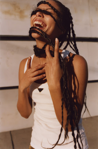 Smiling woman with long braids covering her face, wearing a white tank top against a neutral background, captured in a medium shot.