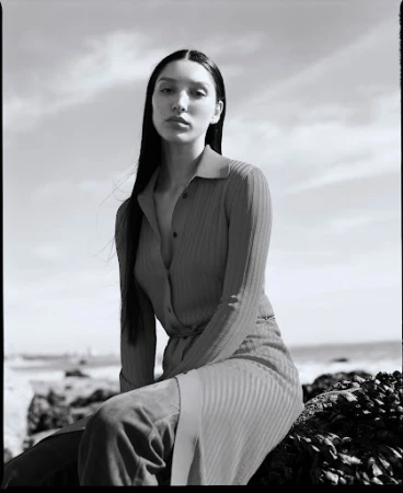 Black and white portrait of a woman with long dark hair wearing a collared button-down top, seated outdoors near a coast.