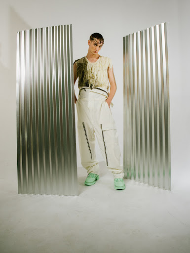 Adult male model wearing a light colored fringed top and trousers in a photo studio setting with metal corrugated panels.