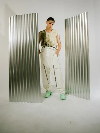 Adult male model wearing a light colored fringed top and trousers in a photo studio setting with metal corrugated panels.