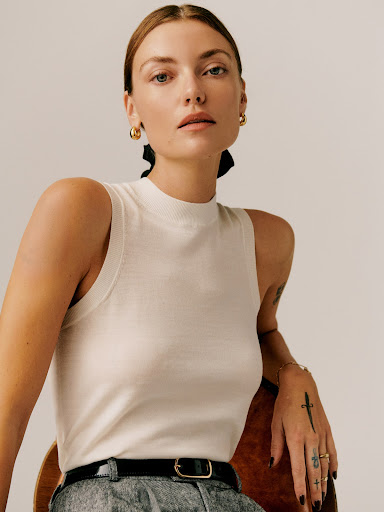 Portrait of a woman in a sleeveless knit top and grey pants. The model wears gold earrings and sits on a wooden chair.