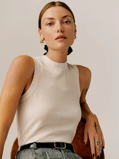 Portrait of a woman in a sleeveless knit top and grey pants. The model wears gold earrings and sits on a wooden chair.