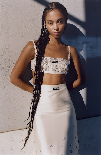 Portrait of a young Black woman with long braids wearing a white embellished crop top and matching skirt against a sunlit wall.