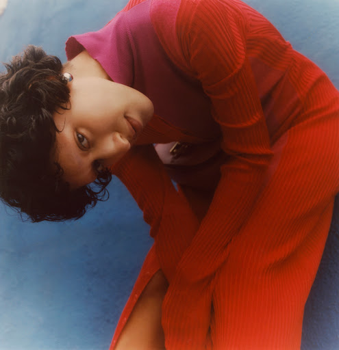 Woman with curly hair poses indoors, wearing red-pink ribbed dress against a blue backdrop, looking down, creative fashion photography.