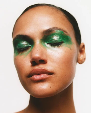 Close-up portrait of a woman with dramatic glossy green eyeshadow makeup across her closed eyes against a plain background