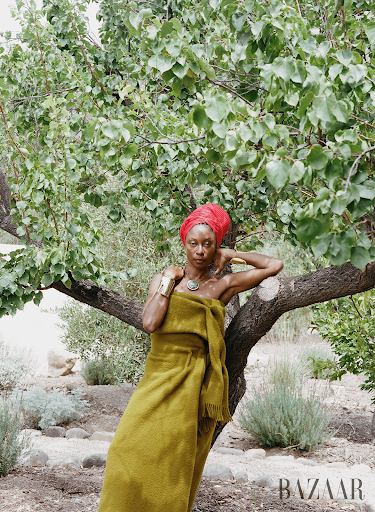 An African American woman wrapped in a fringed chartreuse blanket poses outdoors near a tree in a fashion editorial, with a red head wrap and jewelry.