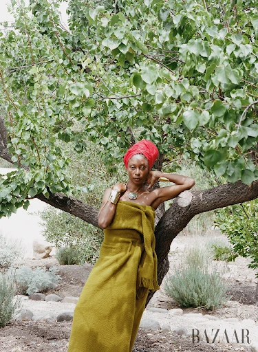 An African American woman wrapped in a fringed chartreuse blanket poses outdoors near a tree in a fashion editorial, with a red head wrap and jewelry.