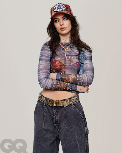 Fashionable woman posing in a graphic long sleeve top, dark denim carpenter pants, studded belt, and baseball cap on a white backdrop.